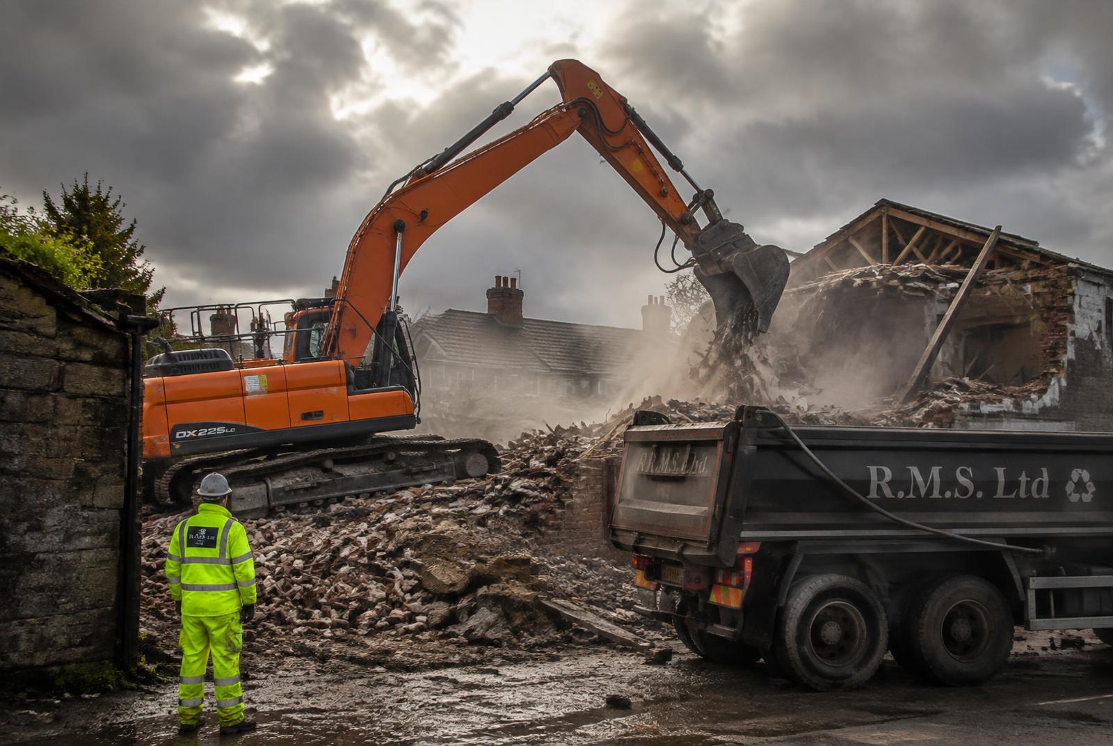 Excavator carrying out controlled demolition of an outbuilding with a muck away lorry alongside